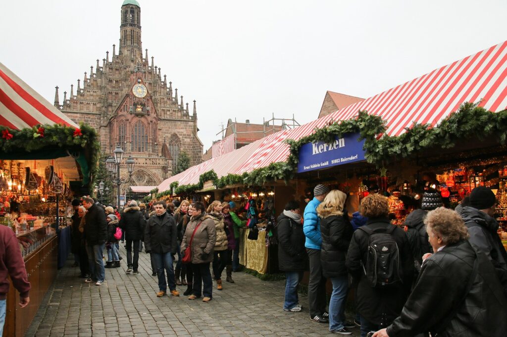 Mercado de Natal em Nuremberg movimentado com várias pessoas caminhando e fazendo compras em barracas decoradas com toldos listrados em vermelho e branco. Ao fundo, destaca-se uma igreja gótica imponente com um grande relógio dourado em sua fachada.