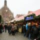 Mercado de Natal em Nuremberg movimentado com várias pessoas caminhando e fazendo compras em barracas decoradas com toldos listrados em vermelho e branco. Ao fundo, destaca-se uma igreja gótica imponente com um grande relógio dourado em sua fachada.